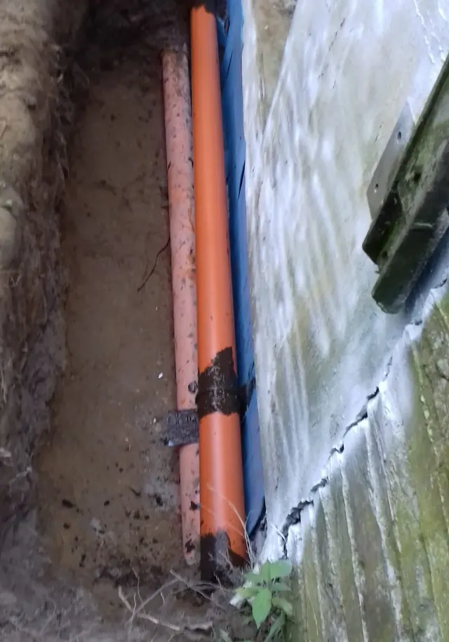 Freigelegte Kellerwand im offenen Graben mit Bitumenabdichtung, blauer Noppenbahn und Drainrohr